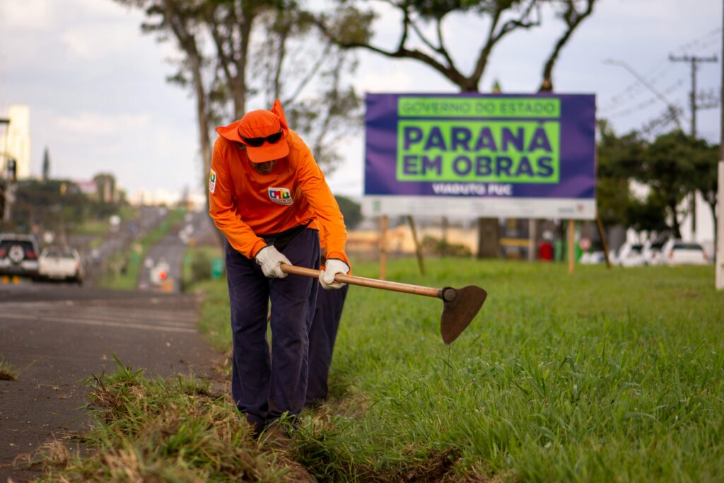 A worker wearing orange uniform maintains roadside grass in Londrina, Brazil.