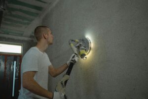 Photo of a man using a power tool to sand a wall during an indoor home renovation project.